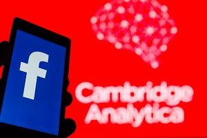  Smartphone in hand with logo of popular social network Facebook. Cambridge Analytica emblem in background.