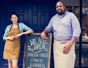 Cheerful restaurant owners standing with "we are open" blackboard.