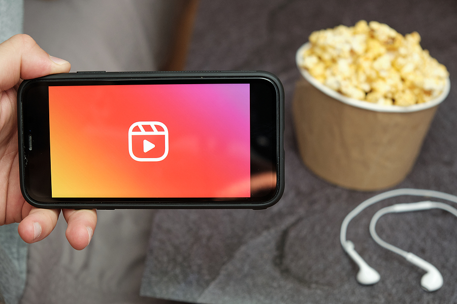 A close up of a man's hand holding a phone with the Instagram Reels logo on the screen over a wooden table, atop of which sits a bowl of popcorn an earbuds.