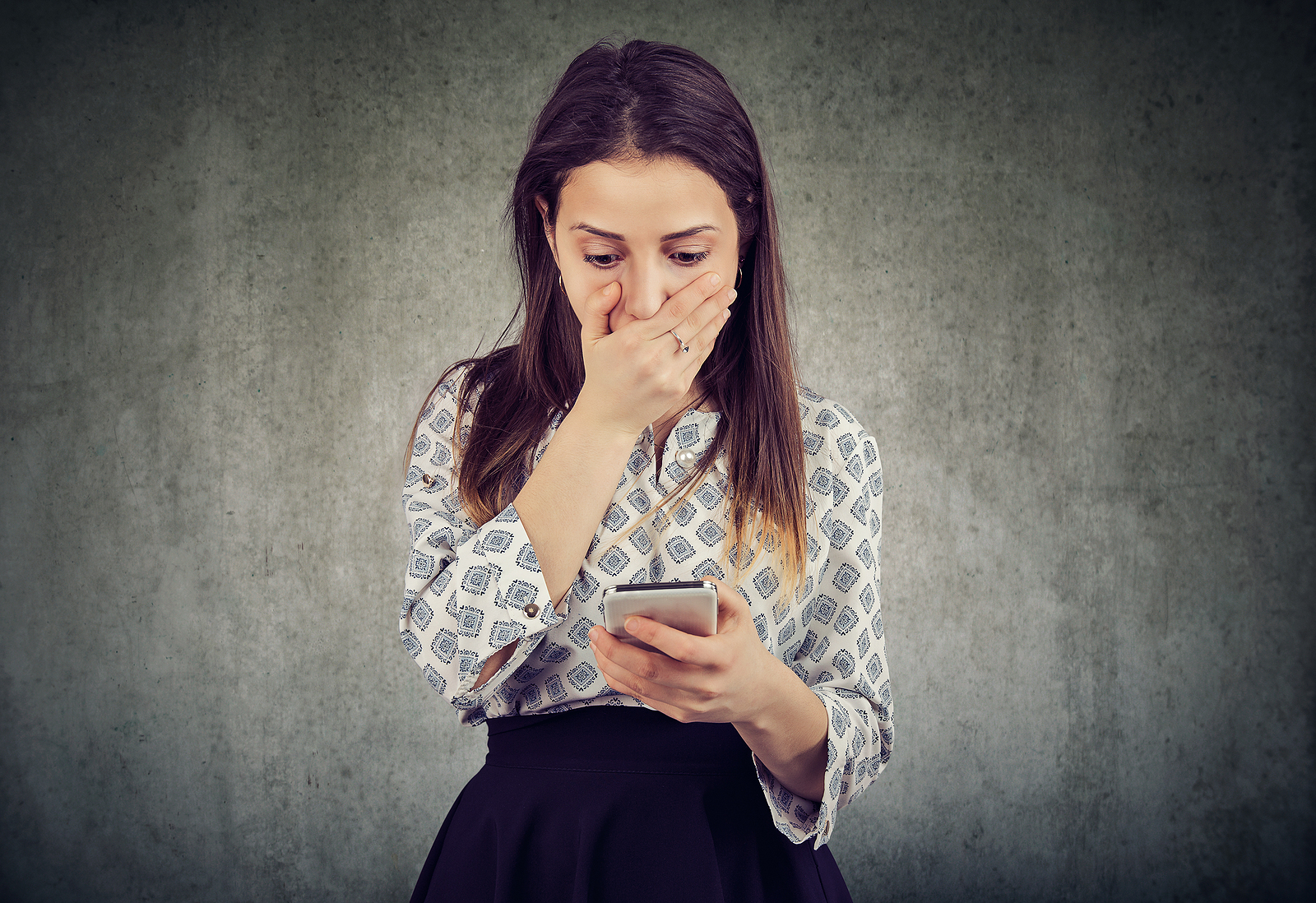 Young woman covering mouth in astonishment while reading message on smartphone looking stunned.