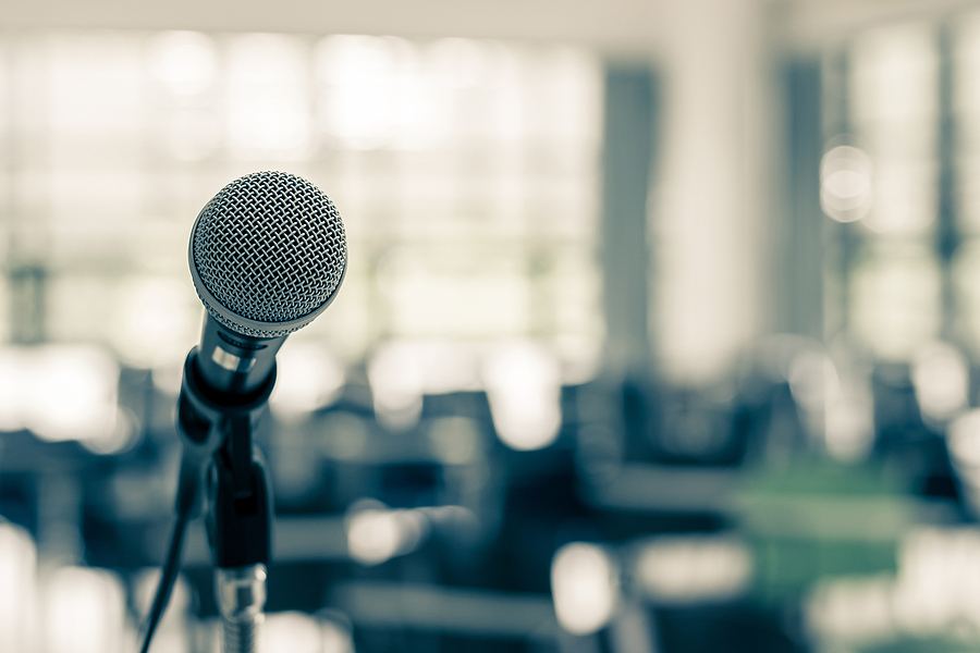 A microphone on a stage with a fuzzy backdrop of an auditorium set up for a seminar